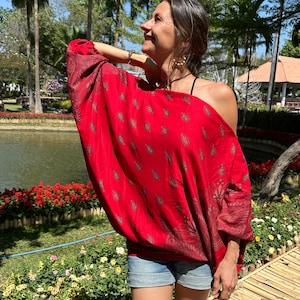 Peut inclure: Une femme portant un haut rouge à épaules dénudées avec un motif paisley et un short en jean. Elle se tient dans un jardin avec un étang et des fleurs.