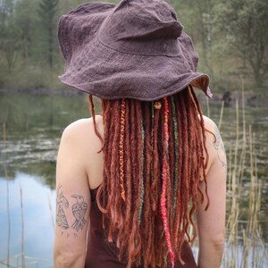 May include: A brown, wide-brimmed hat with a textured appearance. The person is wearing a brown dress and has long, reddish-brown dreadlocks with colorful accents. A tattoo of two birds is visible on the arm.