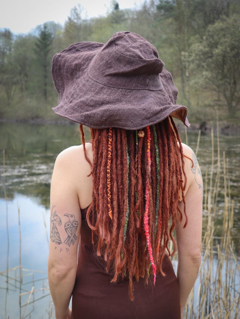 May include: A brown, wide-brimmed hat with a textured appearance. The person is wearing a brown dress and has long, reddish-brown dreadlocks with colorful accents. A tattoo of two birds is visible on the arm.