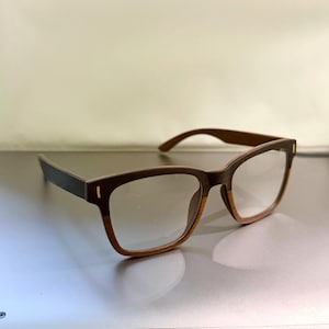 Peut inclure: Lunettes dotées d'une monture bicolore avec une partie supérieure brun foncé et une partie inférieure dans un ton bois naturel plus clair. Les branches sont brun foncé et les verres rectangulaires sont transparents. Texture bois visible.