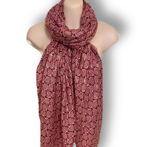 May include: A burgundy scarf with a repeating paisley pattern in cream and gold. The scarf is draped over a mannequin, showcasing its length and design. The fabric appears lightweight and suitable for various seasons.
