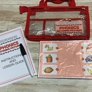 May include: A red zippered tote bag with a clear plastic window containing a set of phonics matching picture cards. The cards are white with red and black text and colorful illustrations. The tote bag also includes a set of instructions and a lesson guide.