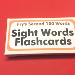 May include: A rectangular box of "Fry's Second 100 Words Sight Words Flashcards" is displayed on a red surface. The box has a white background with an orange border and brown text.