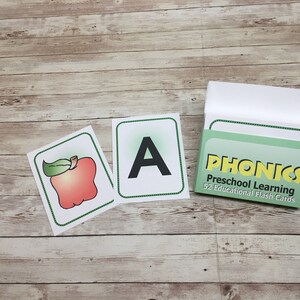 May include: A set of 52 educational flash cards for preschool learning. The cards feature a green background with the word "PHONICS" in yellow letters. The box also says "Preschool Learning" and "52 Educational Flash Cards". Two example cards are shown, one with a red apple and the other with the letter "A" in black.