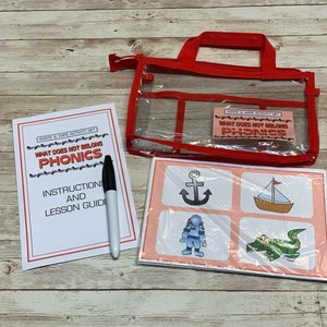 May include: A red and clear plastic bag with a handle contains a set of cards for a "What Does Not Belong Phonics" game. The cards have images of an anchor, a sailboat, an astronaut, and an alligator. A white and red instruction sheet with the title "What Does Not Belong Phonics" and a black dry-erase marker are also included.