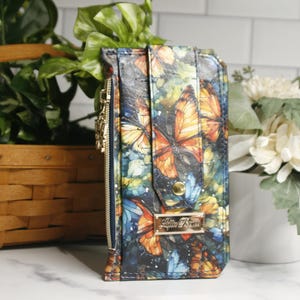 Puede incluir: Una cartera de cuero negro con un estampado de mariposas de colores y una cremallera dorada. La cartera tiene una placa de metal dorada con el texto "Little Fox" en ella.