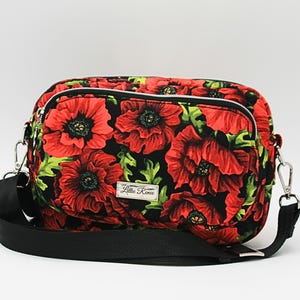 Könnte beinhalten: Schwarze Umhängetasche mit einem auffälligen Blumenmuster aus roten Mohnblumen und grünen Blättern. Sie hat einen verstellbaren schwarzen Riemen, silberfarbene Beschläge und eine Reißverschluss-Vordertasche. Das Etikett lautet "Lillie Renee".