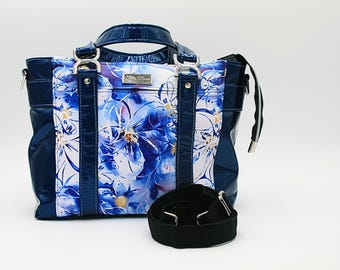 May include: A navy blue tote bag with a floral pattern in shades of blue and white. The bag has a glossy finish, silver hardware, and a black shoulder strap. The handles are navy blue and the bag has a rectangular shape.