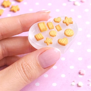 Pode incluir: Um molde de resina transparente em forma de coração preenchido com pingentes em miniatura em forma de bolacha. Os pingentes têm várias formas, incluindo quadrados, retângulos, estrelas e corações. O fundo é um padrão de bolinhas rosa.