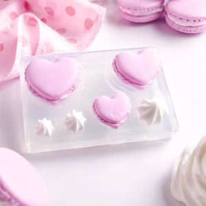 Pode incluir: Um close-up de macarons rosa em forma de coração e merengues brancos. Os macarons estão em um molde transparente, com um pano rosa com bolinhas no fundo. Outros macarons estão espalhados ao redor do molde. A imagem está em um fundo branco.