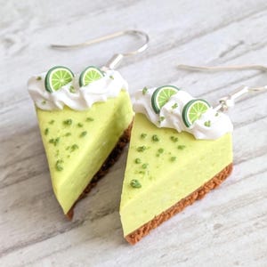 Op de afbeelding: Een paar hangende oorbellen in de vorm van stukjes limoentaart. Elke oorbel heeft een lichtgroene driehoekige vulling, een bruine korstbodem, een toefje witte slagroom en twee kleine groene schijfjes limoen als garnering.