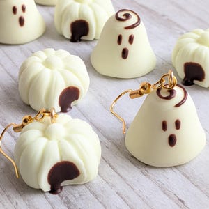 Pode incluir: Um par de brincos com pingentes em forma de fantasma branco com traços faciais castanho-escuros e uma espiral no topo, ao lado de vários pingentes soltos de fantasmas e abóboras brancos com detalhes em castanho-escuro.