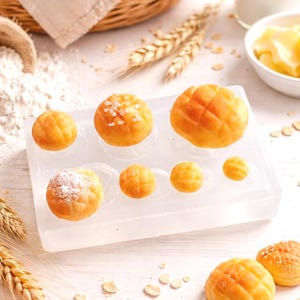 Könnte beinhalten: Ein Stillleben mit Backwaren und Zutaten. Ein Korb mit Croissants und Brot steht neben einem Sack Mehl, einem Holzschöpfer und einer transparenten Form mit kleinen, runden, goldbraunen Backwaren. Butter und Milch sind ebenfalls vorhanden.