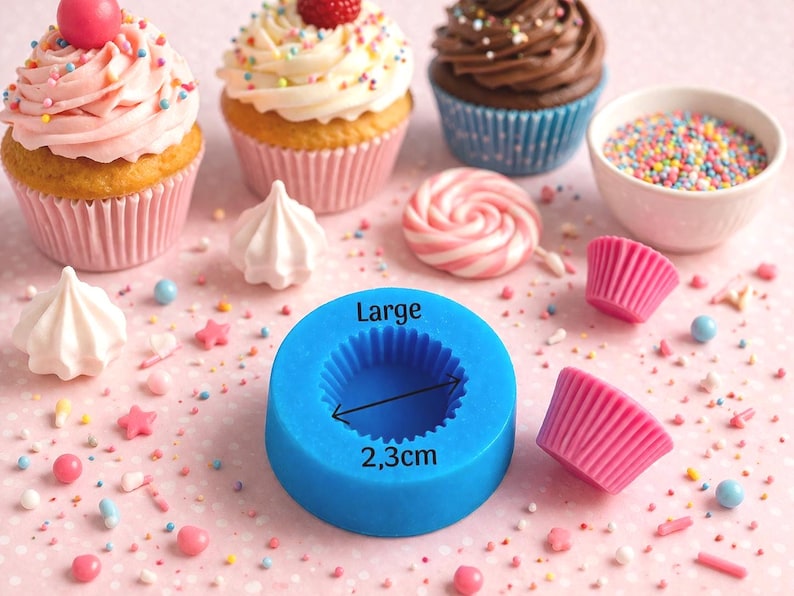 May include: An assortment of cupcakes with colorful frosting and toppings, including sprinkles and a cherry. A blue silicone mold with the text "Large" and a measurement of 2.3cm is also present, along with meringue cookies and a lollipop. The scene is set on a pink surface.