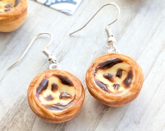 May include: A pair of silver-hook earrings featuring miniature, detailed replicas of Portuguese custard tarts. The earrings have a silver hook and a realistic, baked appearance with a creamy filling and a golden-brown crust. The earrings are on a light-colored wooden surface.