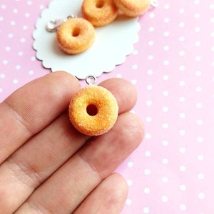 May include: A close-up of a small, yellow, sugared donut charm with a hole in the center. The donut is hanging from a silver chain and is being held by a hand.