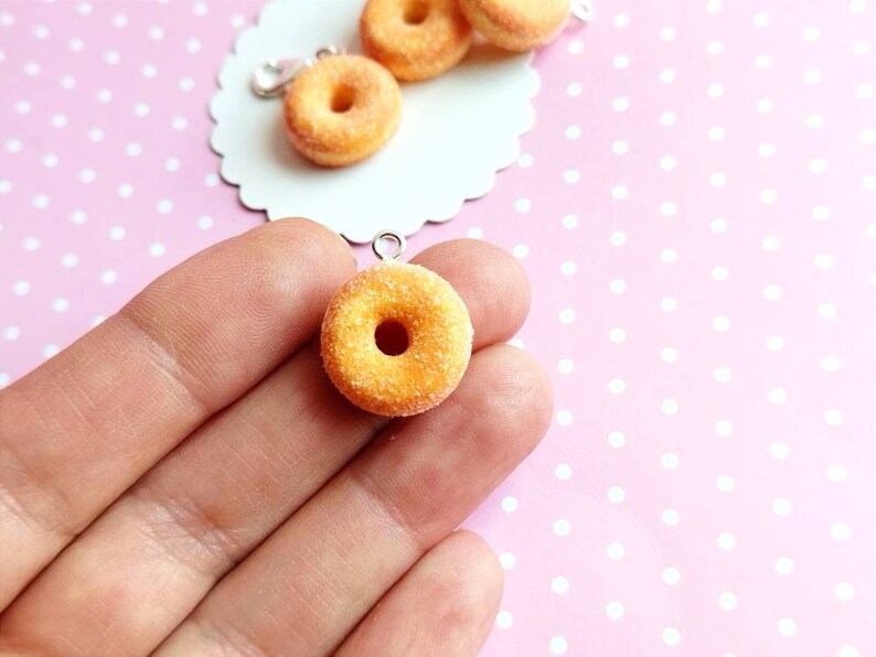 May include: A close-up of a small, yellow, sugared donut charm with a hole in the center. The donut is hanging from a silver chain and is being held by a hand.