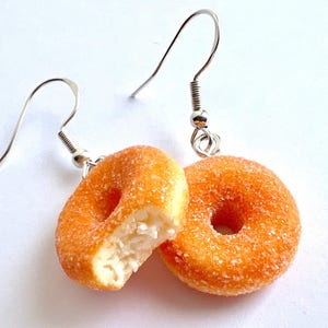 Könnte beinhalten: Ein Paar Donut-Ohrringe. Ein Donut ist ganz und der andere ist ein halber Donut, aus dem ein Stück herausgebissen wurde. Beide Donuts sind mit einem zuckerhaltigen Überzug bedeckt.