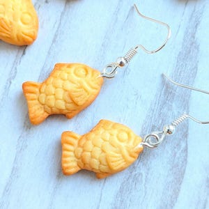 Könnte beinhalten: Ein Paar ausgefallene Ohrringe mit kleinen, goldbraunen fischförmigen Anhängern. Die Anhänger sind an silberfarbenen Ohrhaken befestigt. Mehrere zusätzliche fischförmige Anhänger sind im Hintergrund auf einer hellblauen Oberfläche verstreut.