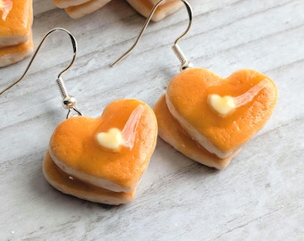 May include: A pair of heart-shaped pancake earrings with a dollop of butter on top. The earrings are made of polymer clay and are a fun and whimsical accessory.