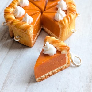Può includere: Un portachiavi a forma di torta di zucca in miniatura con una fetta staccata. La torta ha una crosta marrone dorato e un ripieno arancione vibrante, sormontato da una cucchiaiata di panna montata bianca. Il portachiavi è progettato per assomigliare a un dessert realistico.