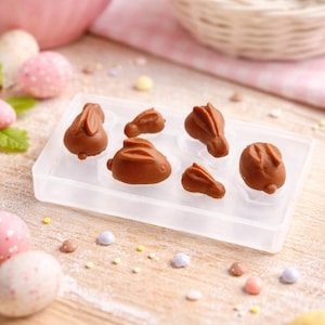 Pode incluir: Um molde de plástico transparente cheio de seis pequenos coelhos de chocolate castanho. O molde está sobre uma superfície de madeira salpicada com doces de ovo da Páscoa em cores pastel. No fundo, há tigelas e cestas cheias de mais ovos da Páscoa.