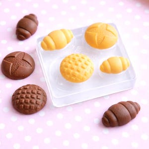 May include: An assortment of miniature bread and pastry shapes in shades of brown and yellow. The collection includes croissants, round buns, and a waffle-like bread, arranged on a pink polka dot surface.