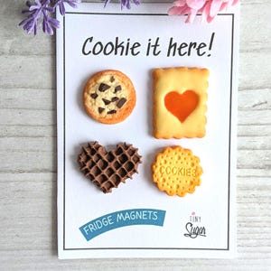 Pode incluir: Um cartão branco com o texto "Cookie it here!" apresenta quatro ímanes de frigorífico em forma de biscoitos. Os ímanes incluem um biscoito com pepitas de chocolate, um waffle em forma de coração e um biscoito com a palavra "COOKIES". O cartão também diz "FRIDGE MAGNETS".