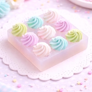 May include: A square mold filled with nine pastel-colored swirls, in shades of pink, blue, green, and white. The mold sits on a white, scalloped doily, with scattered sprinkles in the background. The overall theme is sweet and delicate.