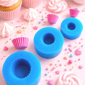 May include: A collection of cupcakes with pink frosting and sprinkles, alongside blue silicone molds, pink cupcake liners, and candies. The scene is set on a pink surface, creating a colorful and festive display.
