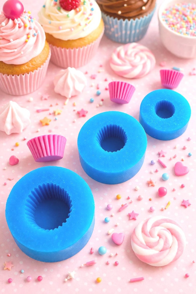 May include: A collection of cupcakes with pink frosting and sprinkles, alongside blue silicone molds, pink cupcake liners, and candies. The scene is set on a pink surface, creating a colorful and festive display.