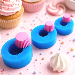 Pode incluir: Uma foto em close-up de cupcakes, doces e moldes de silicone. Os cupcakes têm cobertura rosa e granulado. Moldes de silicone azuis com formas de forminhas de cupcake rosa estão em primeiro plano. O fundo é uma superfície rosa com granulado espalhado.
