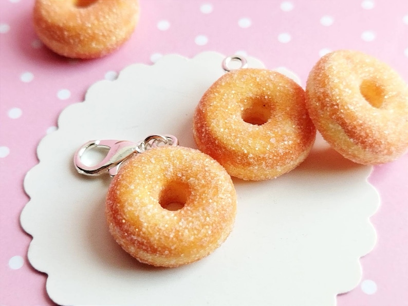 May include: Four sugared mini donuts with a silver charm attached to one. The donuts are on a white plate with a scalloped edge. The plate is on a pink background with white polka dots.