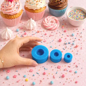 May include: An assortment of decorated cupcakes with pink frosting, sprinkles, and a cherry, alongside blue silicone molds. The cupcakes are in pink and blue paper liners. Colorful sprinkles and candies are scattered on a pink dotted surface.