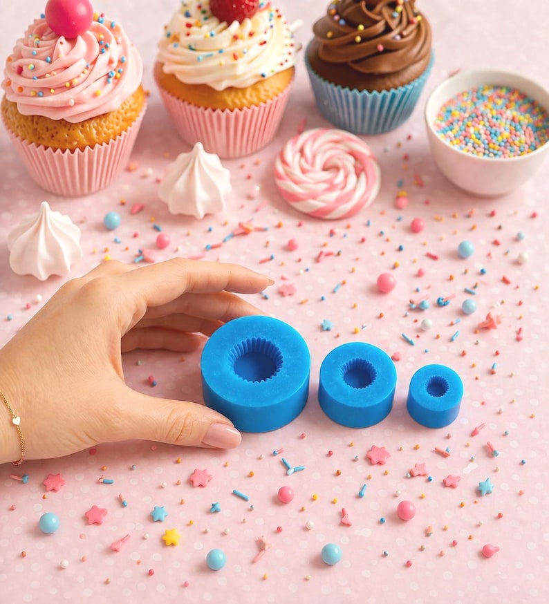 May include: An assortment of decorated cupcakes with pink frosting, sprinkles, and a cherry, alongside blue silicone molds. The cupcakes are in pink and blue paper liners. Colorful sprinkles and candies are scattered on a pink dotted surface.