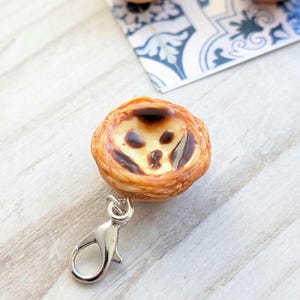 Pode incluir: Um porta-chaves em miniatura de pastel de nata com fecho prateado. A tarte tem uma crosta dourada e estaladiça e um recheio cremoso de creme com um topo ligeiramente queimado. Tartes adicionais são visíveis no fundo sobre uma superfície estampada em azul e branco.