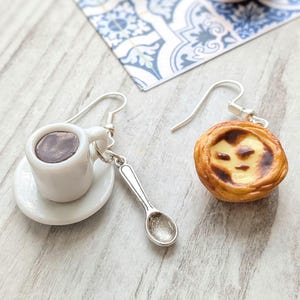 May include: A pair of unique earrings featuring a miniature white coffee cup with a brown liquid, a silver spoon, and a pastry. The pastry has a golden-brown crust and a creamy yellow filling. The earrings are on a light wood surface.
