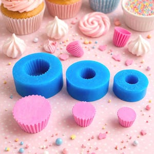 May include: An assortment of cupcakes with colorful frosting and sprinkles, alongside blue silicone molds and pink cupcake liners. Decorative meringues and sprinkles are scattered on a pink polka dot surface.