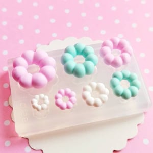 以下が含まれることがあります： パステルカラーのピンク、ミントグリーン、白のドーナツ型レジンパーツが様々なサイズで入った透明シリコンモールド。ピンクのドット柄の背景に置かれています。