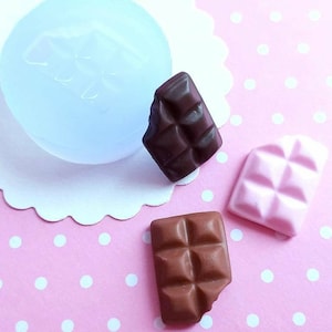 Può includere: Tre mini barrette di cioccolato, una marrone, una rosa e una con un motivo marrone e bianco, su uno sfondo rosa con pois bianchi.