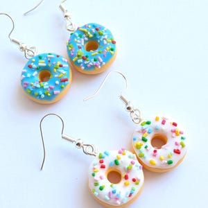 Könnte beinhalten: Ohrringe in Donut-Form mit silbernen Haken. Zwei Paar Ohrringe sind zu sehen, eines mit blauer Glasur und bunten Streuseln, das andere mit weißer Glasur und bunten Streuseln. Die Donuts haben eine hellbraune Basis.