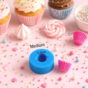 May include: A blue silicone mold, labeled "Medium" with a 1.5cm diameter, sits among cupcakes, meringues, sprinkles, and pink cupcake liners. The scene is set on a pink surface, suggesting a baking or dessert theme.