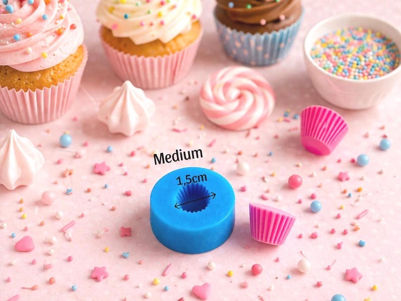 May include: A blue silicone mold, labeled "Medium" with a 1.5cm diameter, sits among cupcakes, meringues, sprinkles, and pink cupcake liners. The scene is set on a pink surface, suggesting a baking or dessert theme.