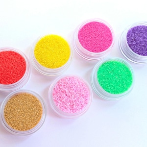 Può includere: Sette piccoli vasetti di plastica trasparente riempiti con sostanze granulari colorate. I colori includono rosso, giallo, rosa, viola, verde, rosa chiaro e oro. I vasetti sono disposti in un motivo circolare su uno sfondo bianco, suggerendo un prodotto artigianale o cosmetico.