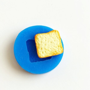 Può includere: Stampo in silicone blu per fare pane in miniatura. Lo stampo ha una forma quadrata con un bordo rialzato. Un piccolo pezzo di pane quadrato è appoggiato sullo stampo.