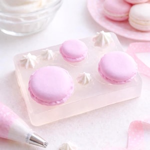 May include: A still life composition featuring pink macarons, white meringue, and a pastry bag. The macarons are displayed on a clear tray and a pink plate. A ribbon with white polka dots adds a decorative touch. The scene is set on a white surface.