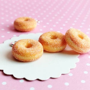 May include: Four miniature sugar-coated donuts with a hole in the center, on a white scalloped surface with a pink polka dot background.