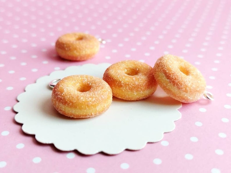May include: Four miniature sugar-coated donuts with a hole in the center, on a white scalloped surface with a pink polka dot background.