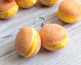 May include: A pair of novelty earrings shaped like miniature peach-colored sandwich cookies with yellow filling. The earrings have a textured, sugary appearance and are suspended from silver-colored ear wires. Other similar earrings are visible in the background.
