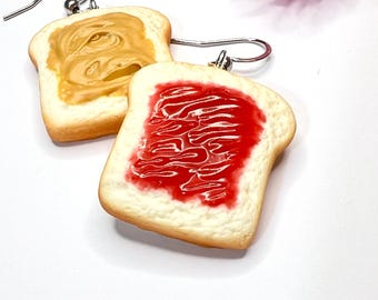 May include: A pair of novelty earrings shaped like slices of toast. One slice is topped with peanut butter, the other with red jelly. The earrings have silver-colored ear wires.
